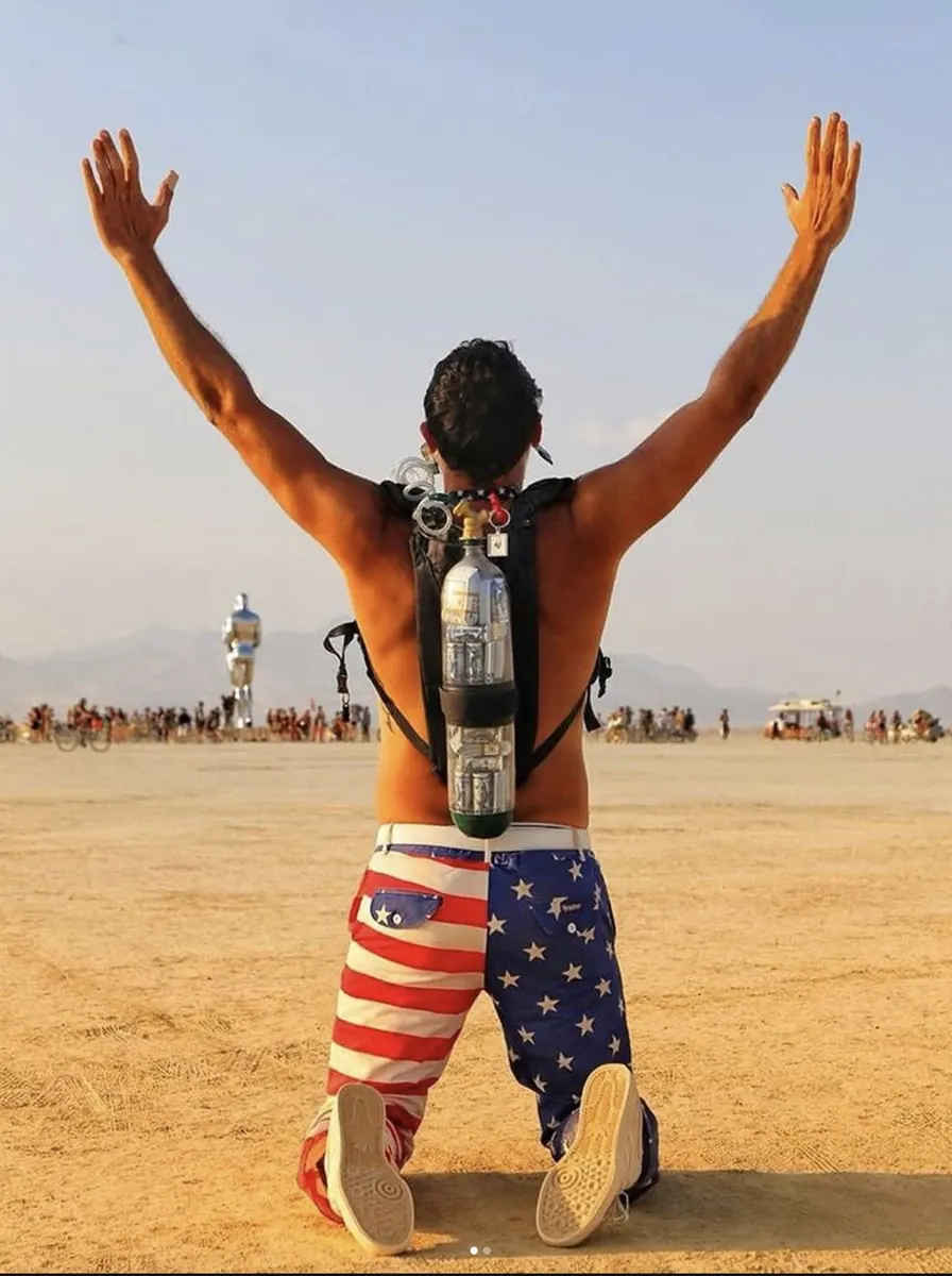 Capitalist Man — kneeling on the playa, tank on back